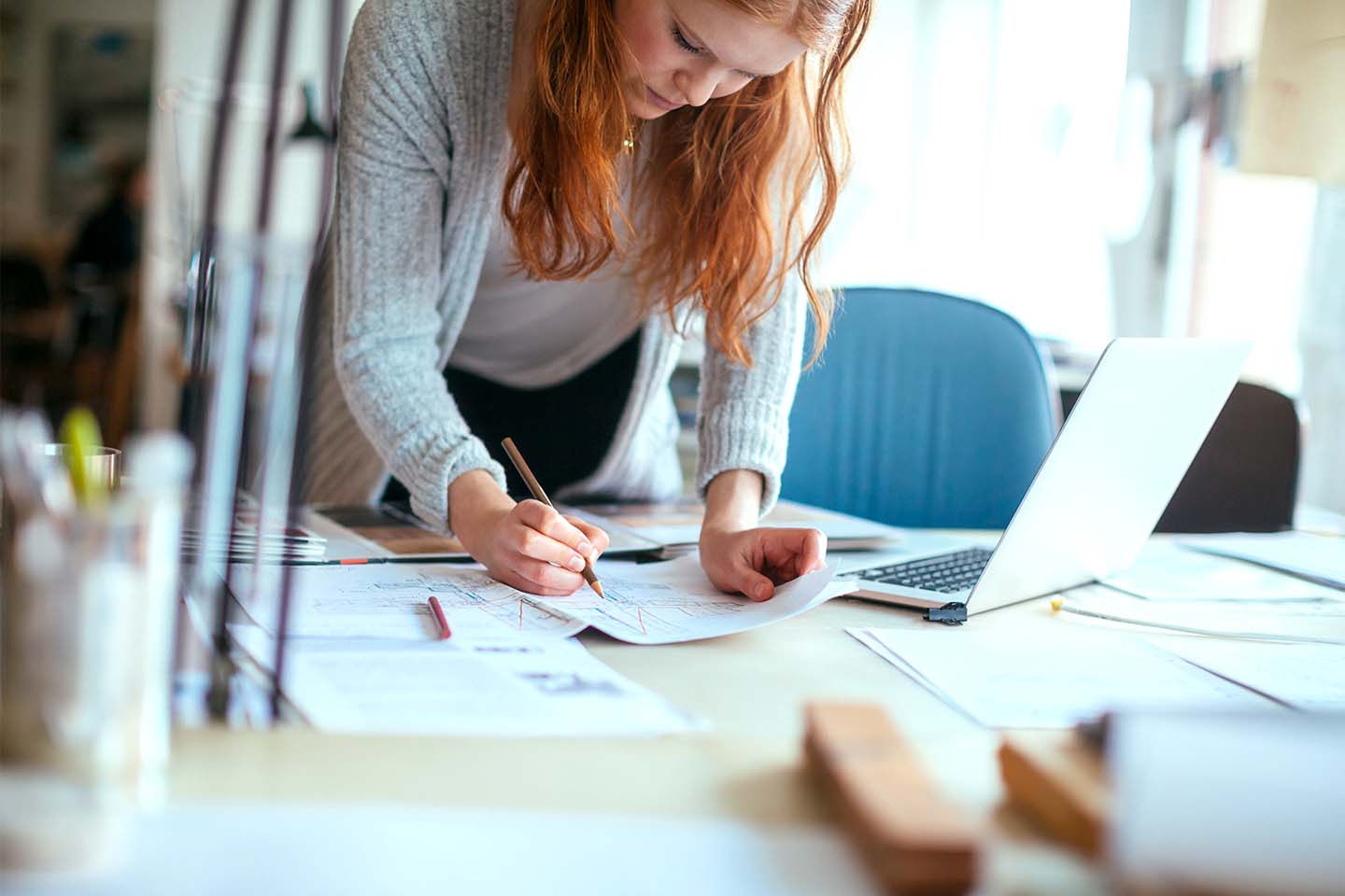 Eine junge Frau mit langen roten Haaren beugt sich über ihren Schreibtisch. Vor ihr befinden sich mehrere große Blätter mit Diagrammen, neben ihr steht ein moderner Laptop. Sie zeichnet mit Bleistift auf einen der Bögen.